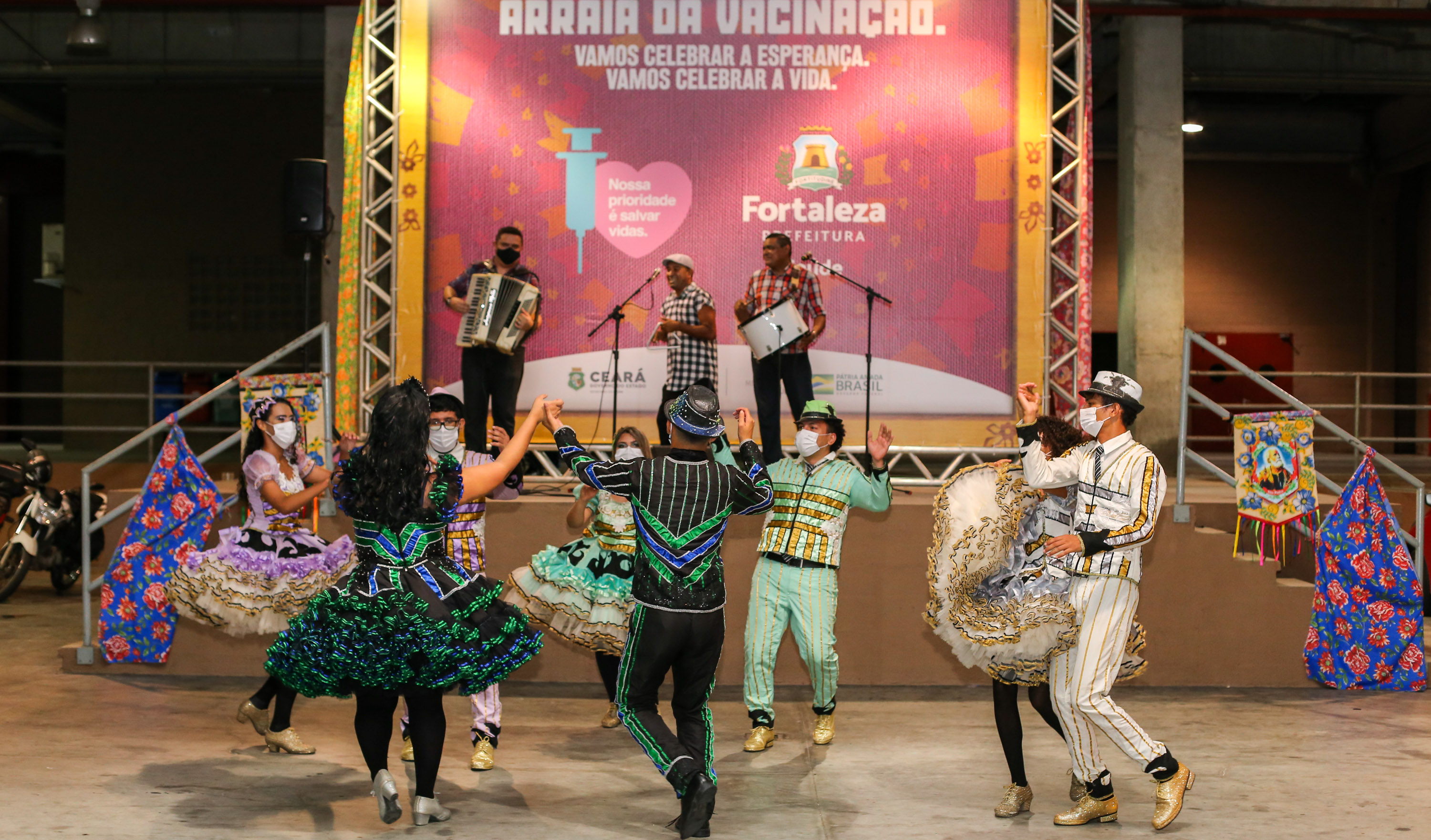 a foto mostra uma grupo de são joão dançando quadrilha e ao fundo um trio com cantor sanfoneiro e tocador de zabumba